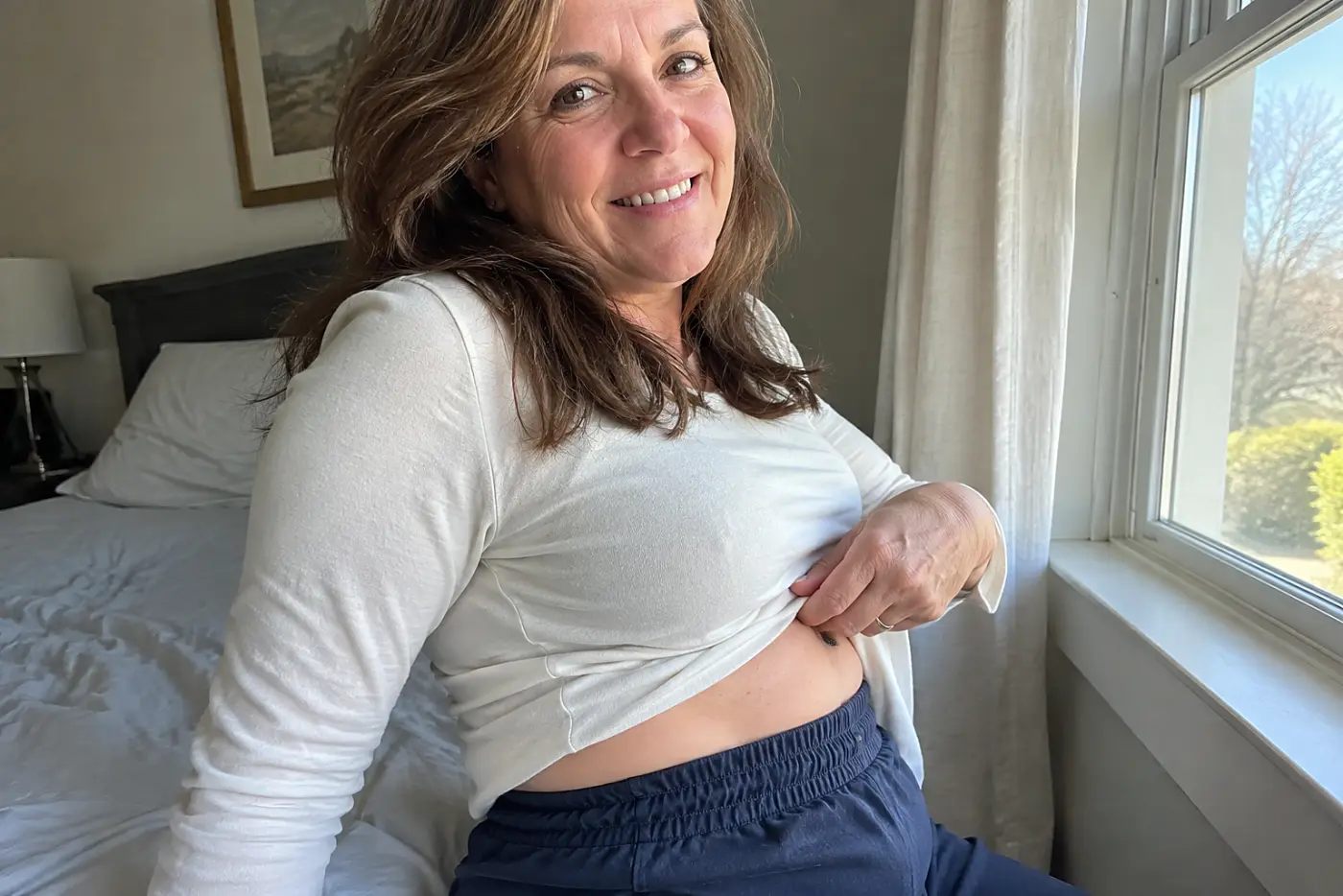 A waistband with side elastic is centered on a woman over 50 seated by a bedroom window, her untucked shirt lifted just enough to show the sides.