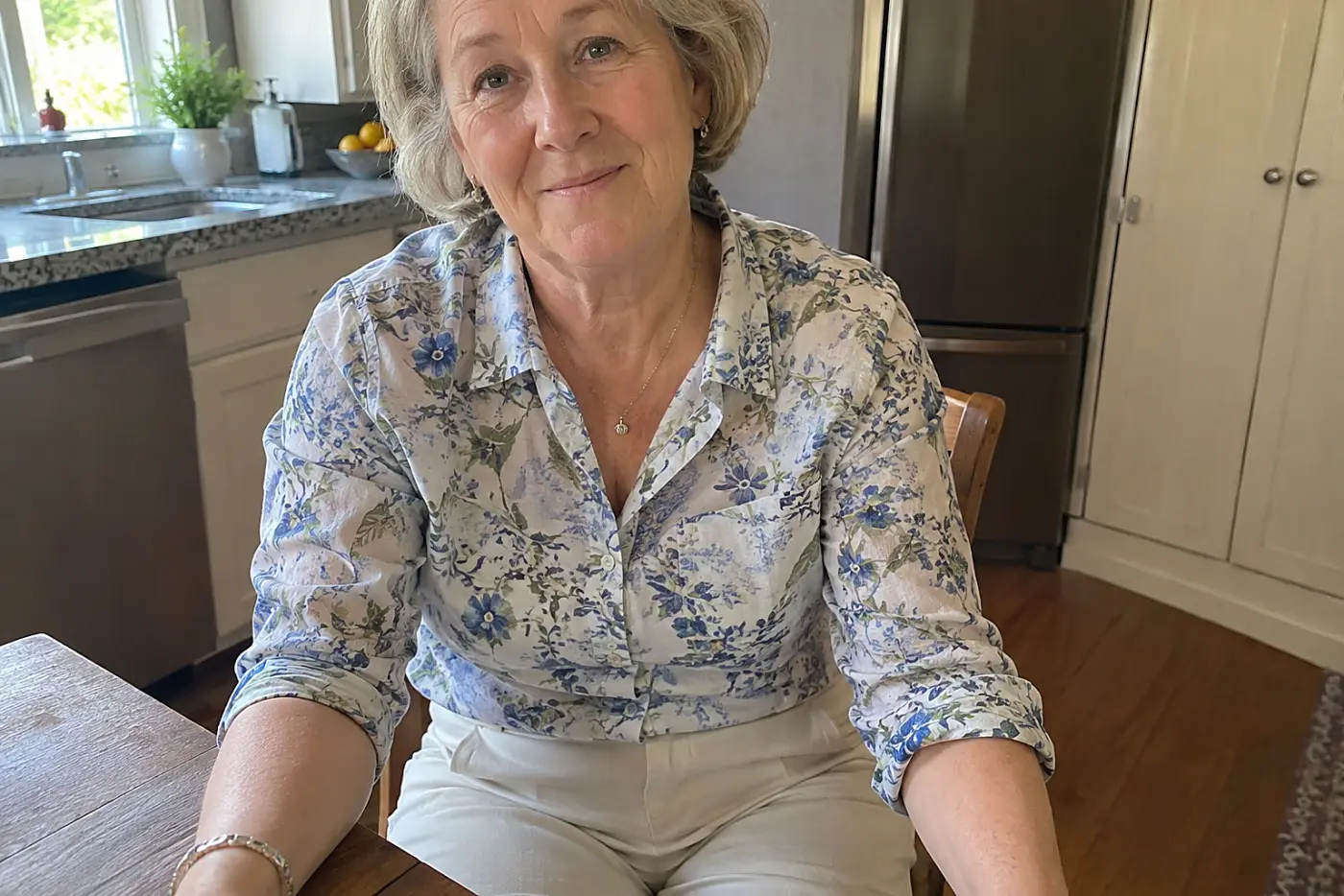 Flat-front pants are centered on a woman in her sixties seated at her kitchen table, with the front waistband visible beneath her shirt.