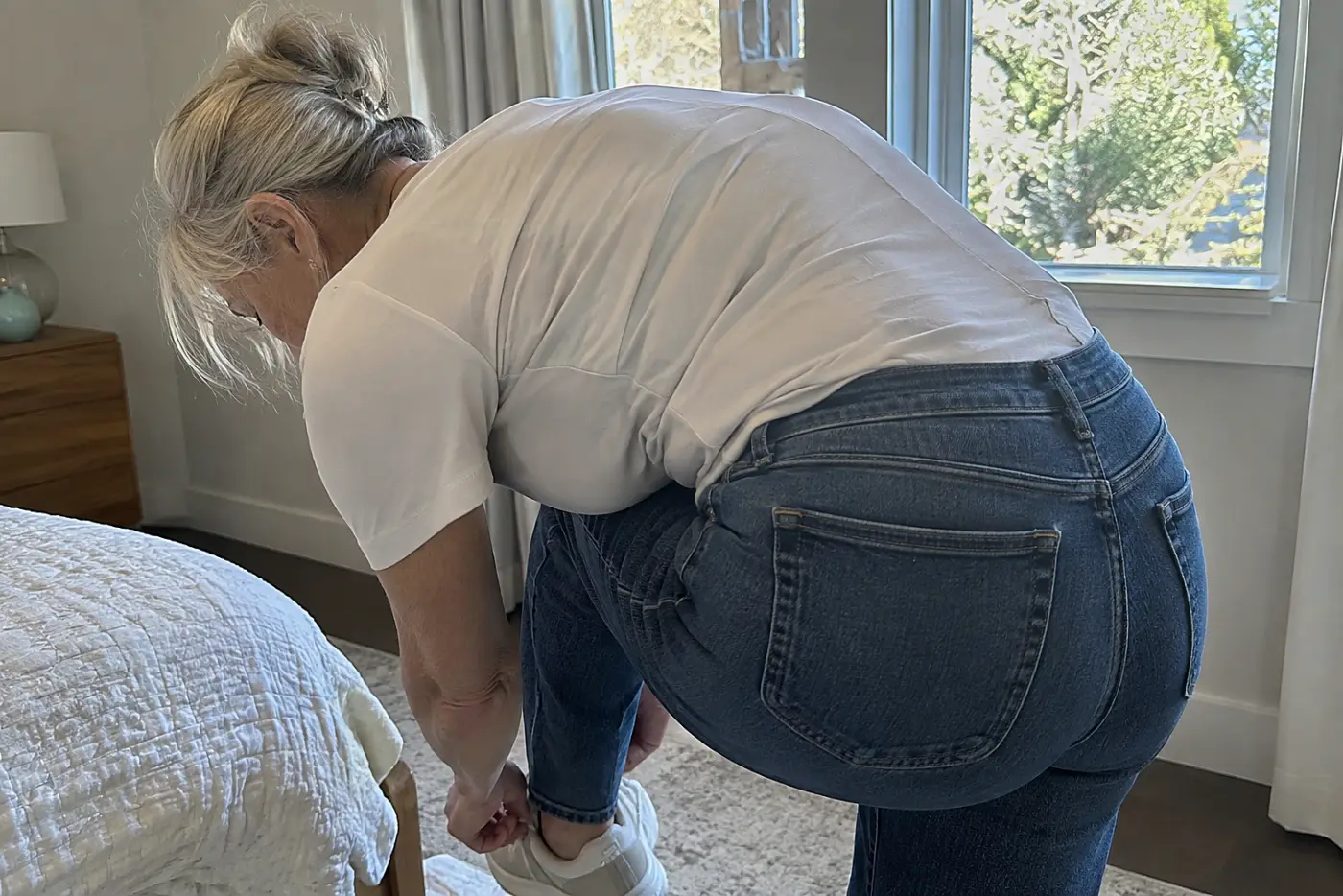 Jeans with a higher back rise sit at the center of the frame as a woman in her sixties bends to tie a shoe in her bedroom.