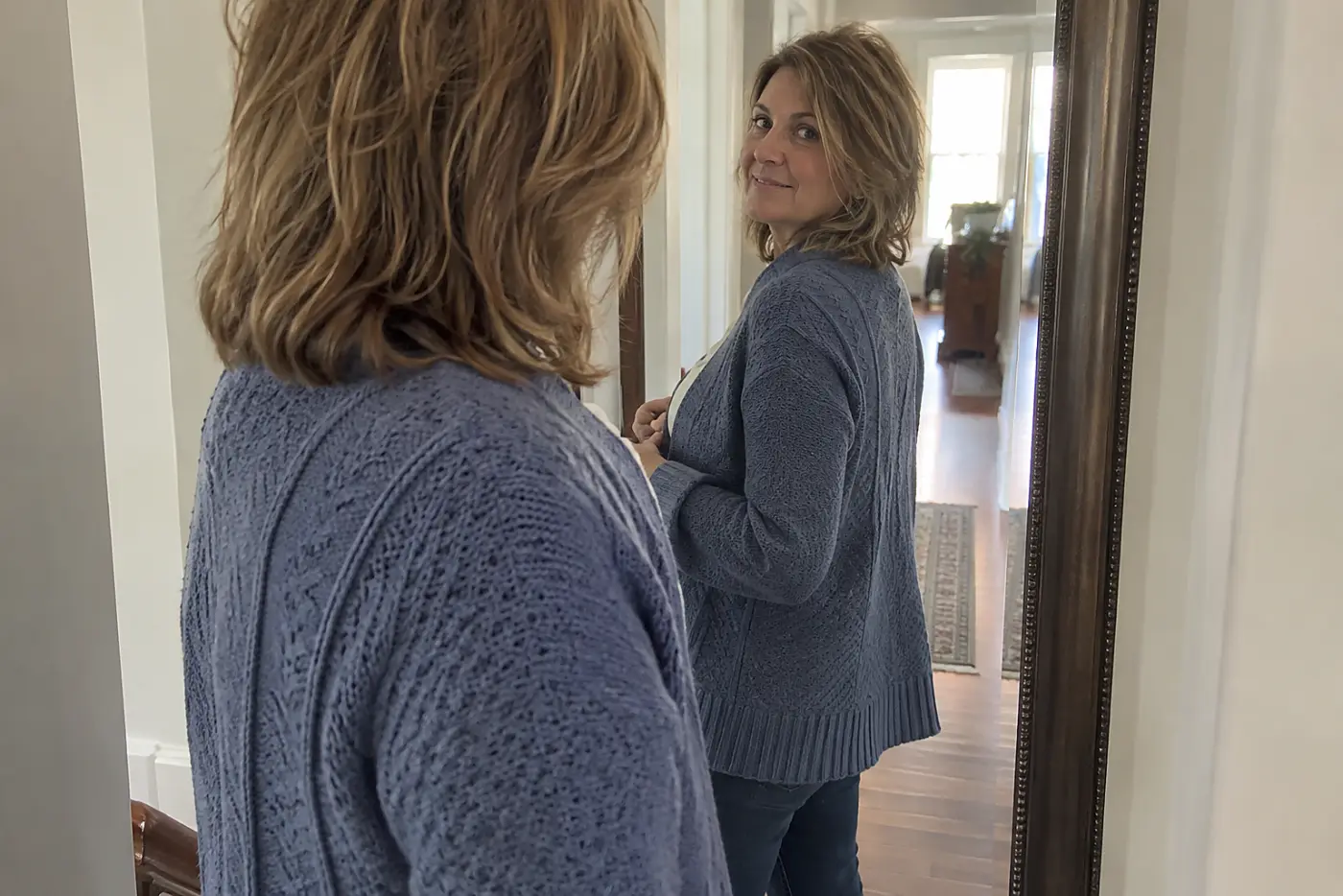 A hip-length cardigan is centered on a woman over 50 turning toward a hallway mirror with the back hem visible.