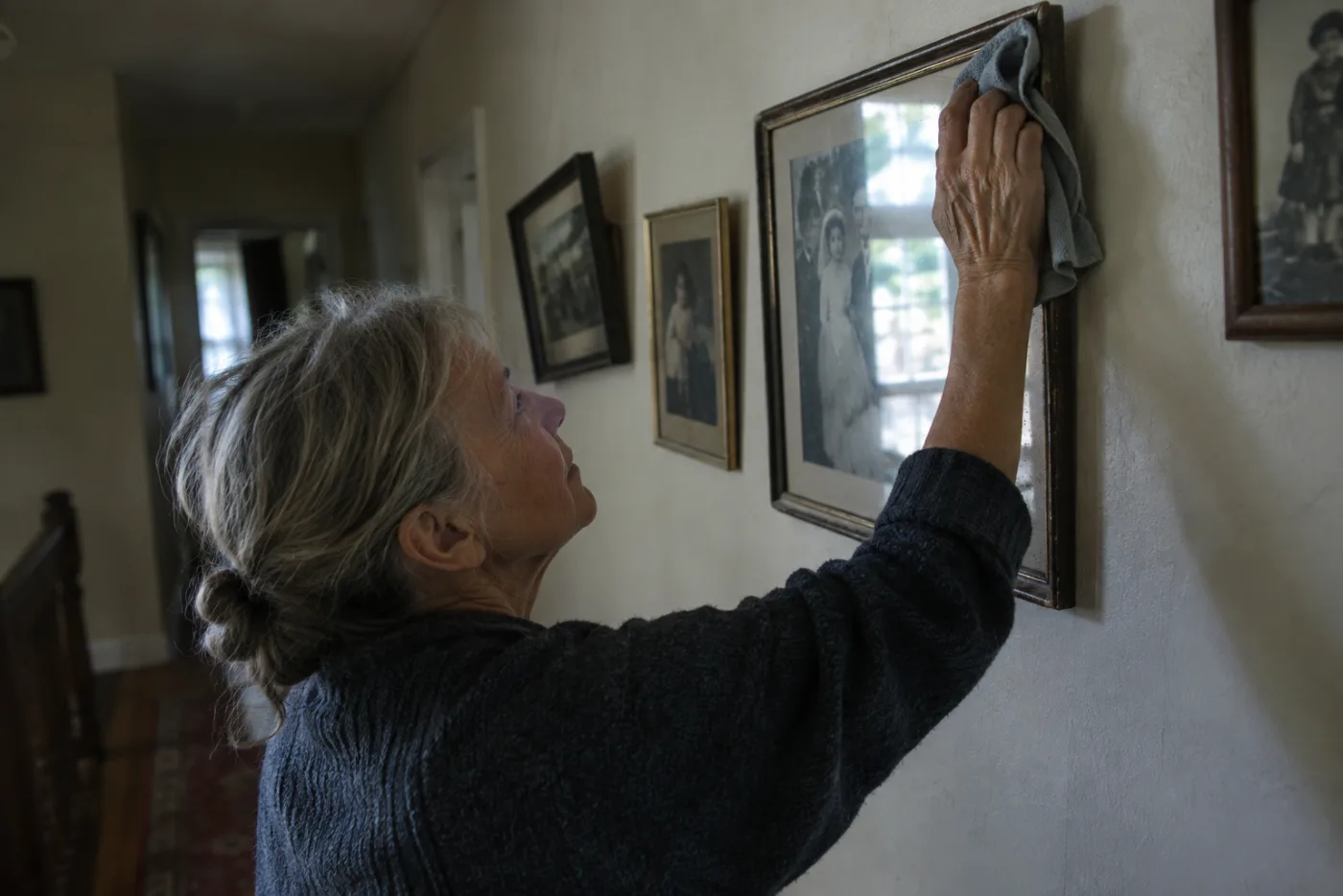 Woman dusting a crooked framed photo in a hallway lined with family pictures