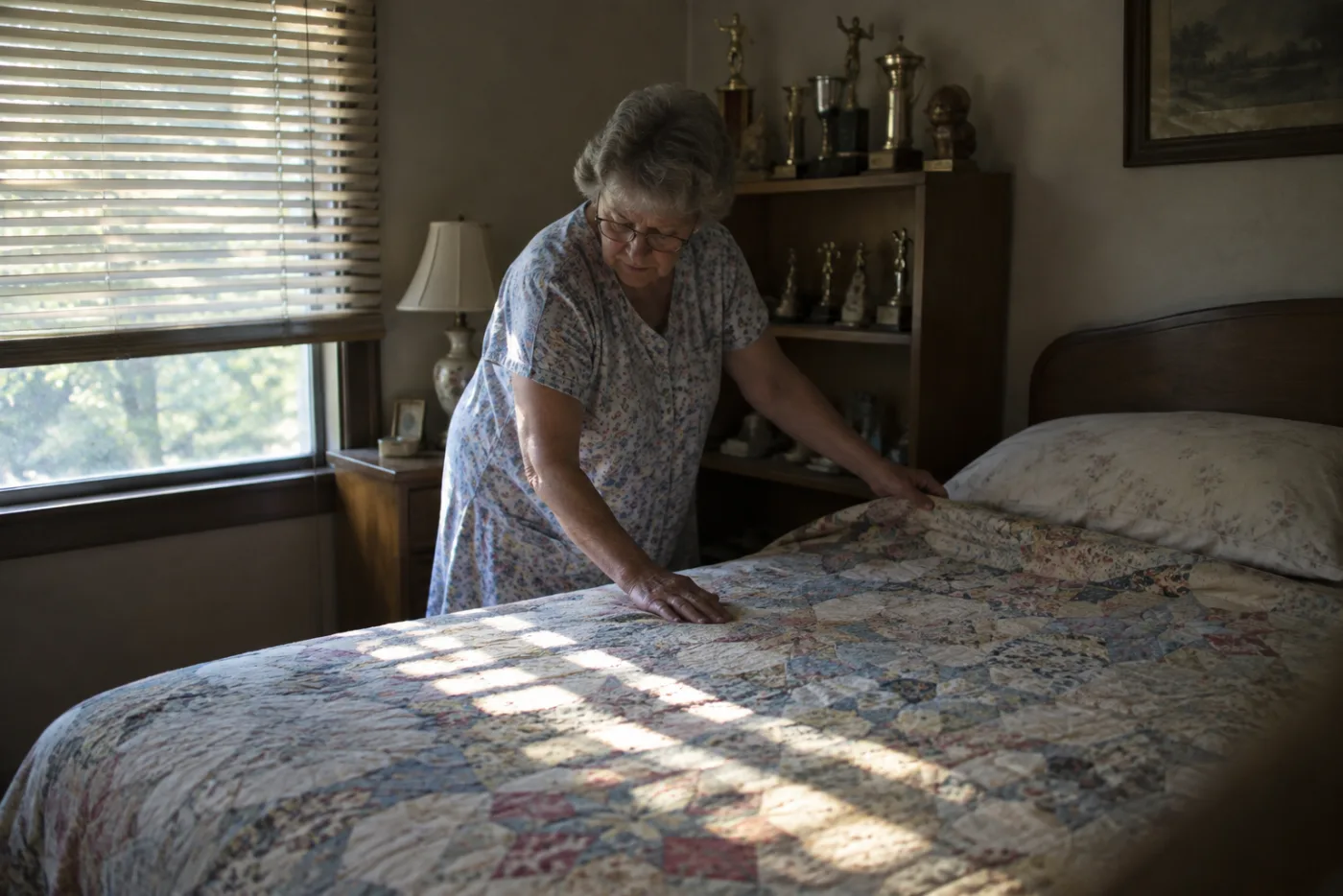Woman smoothing a quilt in an untouched bedroom beside a shelf of dusty trophies