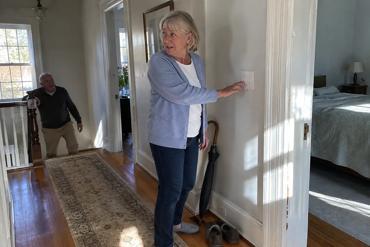 Older woman reaching for a hallway light switch outside a vacant bedroom.