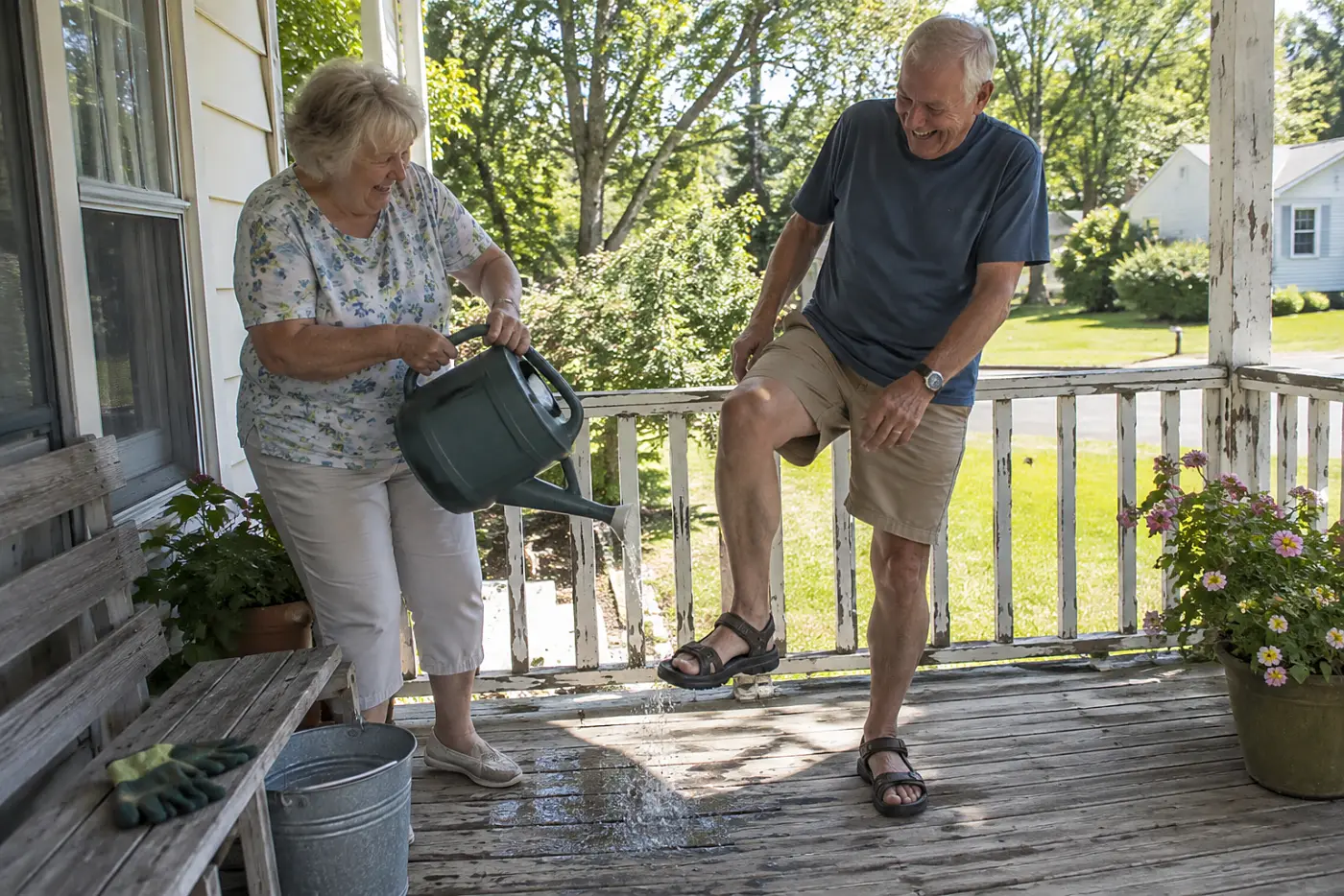 Older woman turning a watering can upside down on a porch bench.