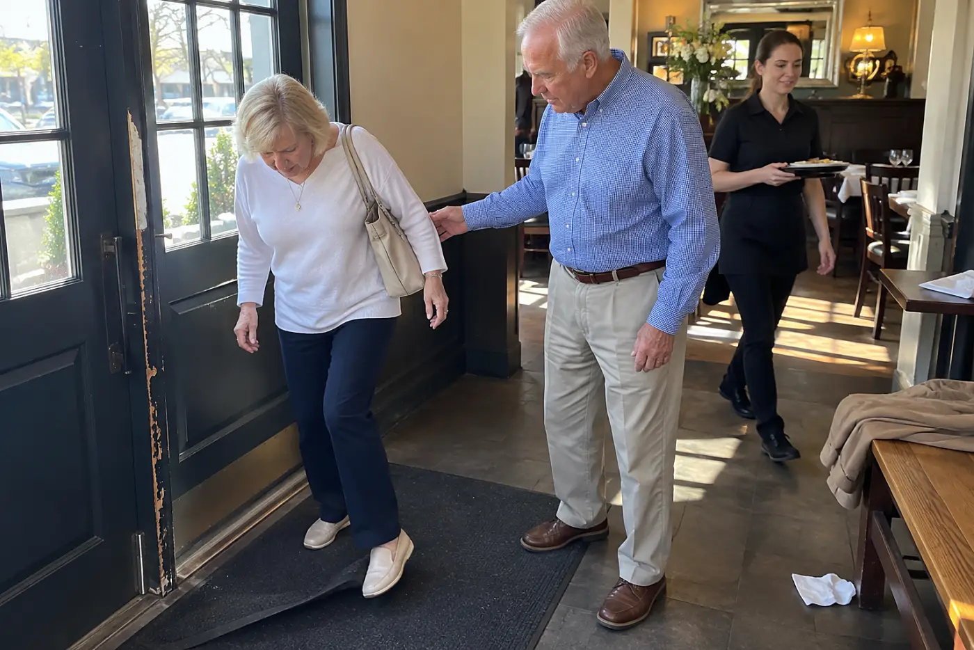 Older woman looking down at a curled mat inside a restaurant entrance.