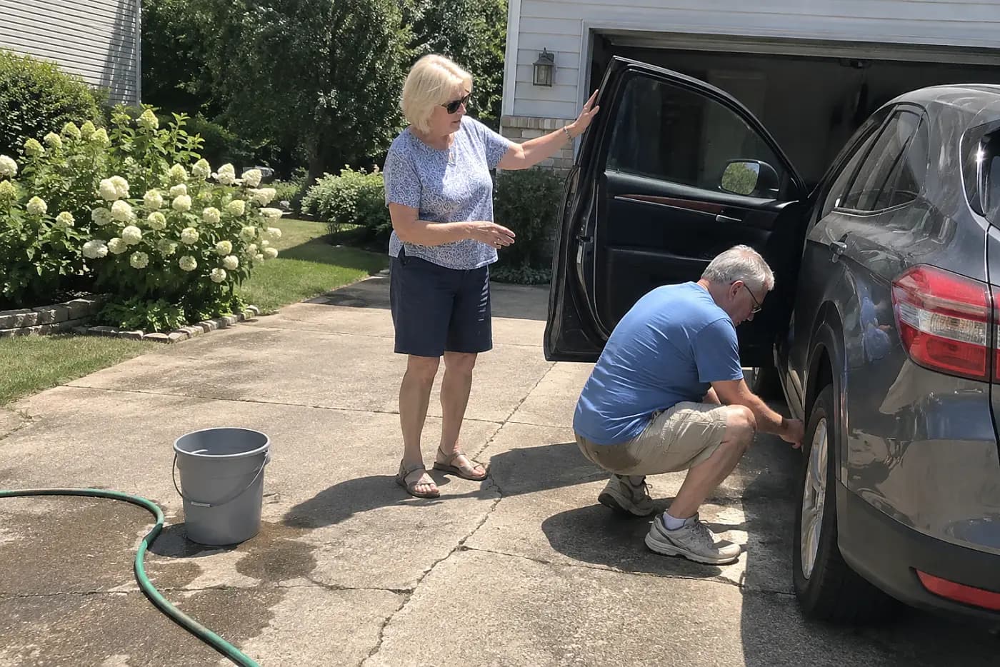 Older woman speaking with her husband beside their parked car in the driveway.