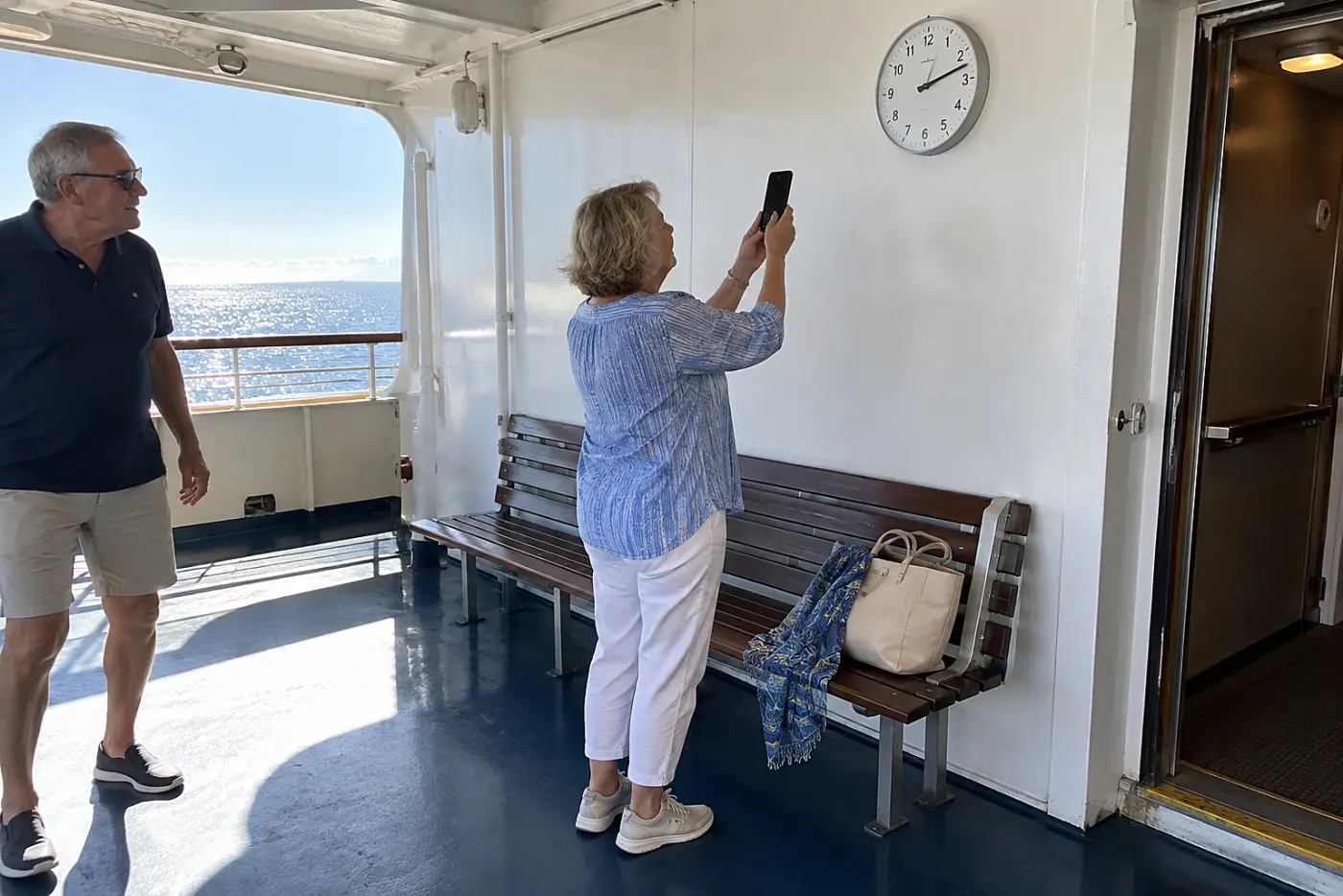 Older woman photographing a clock beside a ship's exit.