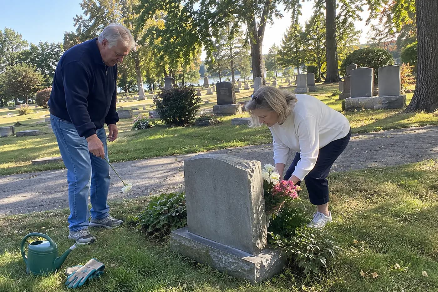 Older woman reading a grave marker along a cemetery path.