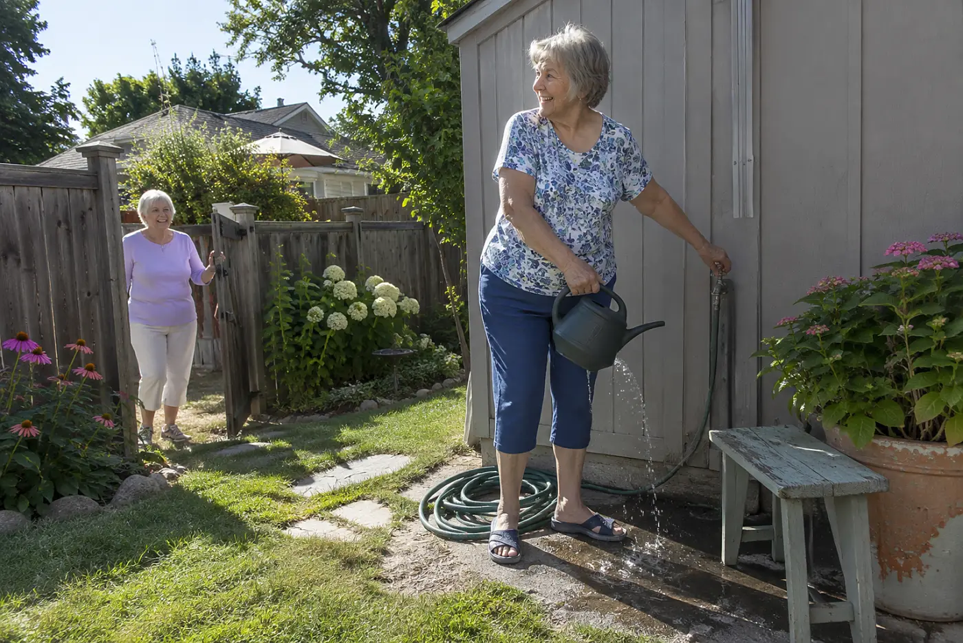 Older woman filling a small watering can halfway at a backyard faucet.