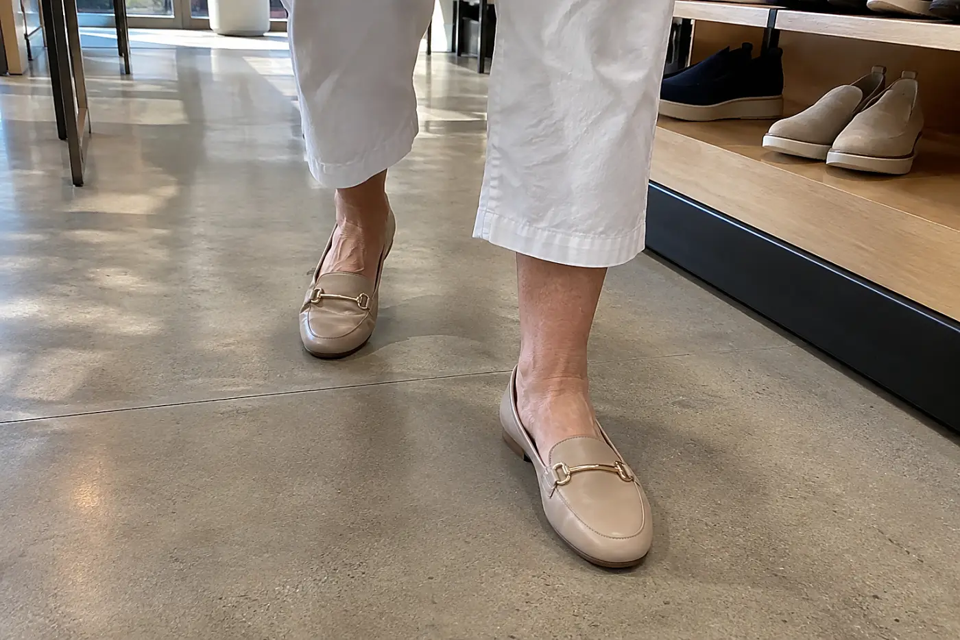A properly fitted shoe is centered on the foot of a woman in her sixties walking naturally across a shoe store's bare floor, with the matching shoe fully visible.