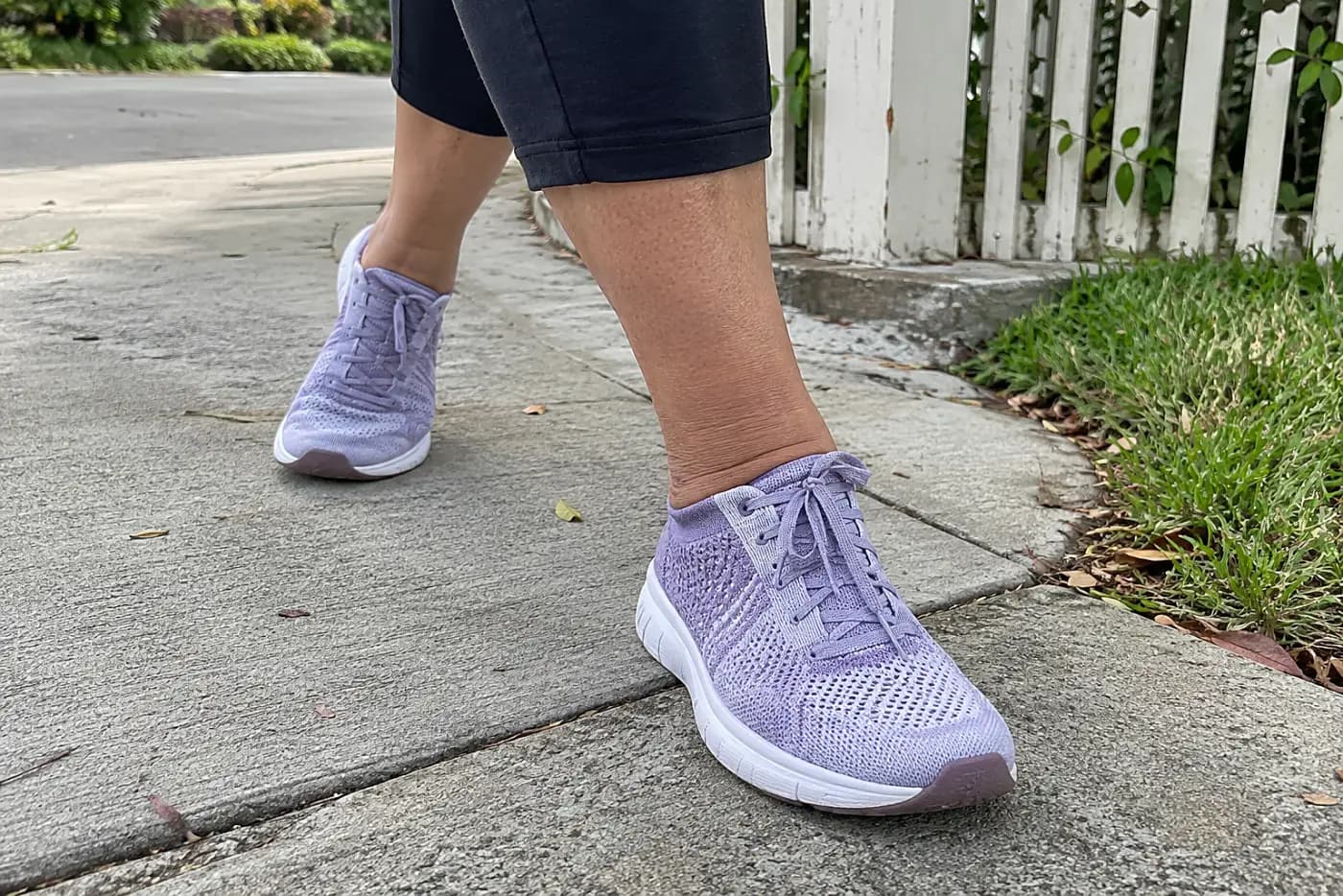 A knit walking sneaker is centered on the foot of a woman over 50 turning a corner on a neighborhood sidewalk.