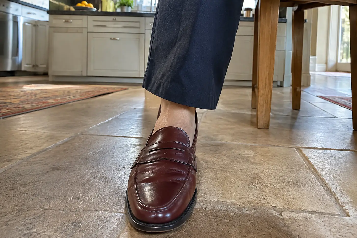 A penny loafer in oxblood is centered beneath the navy trouser leg of a woman over 50 sitting at a kitchen table.
