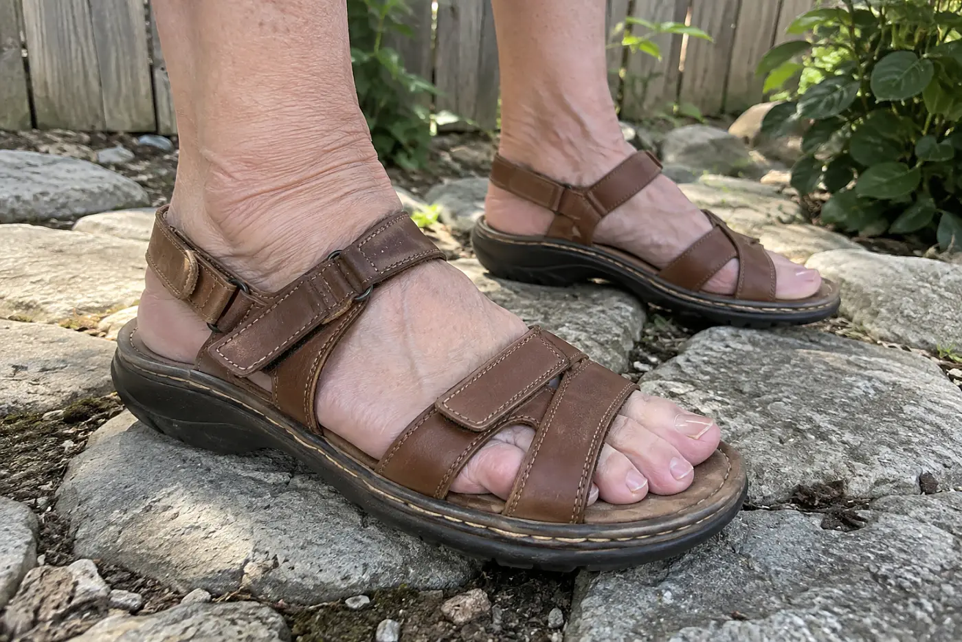A leather walking sandal is centered on the foot of a woman in her sixties standing on a garden path.