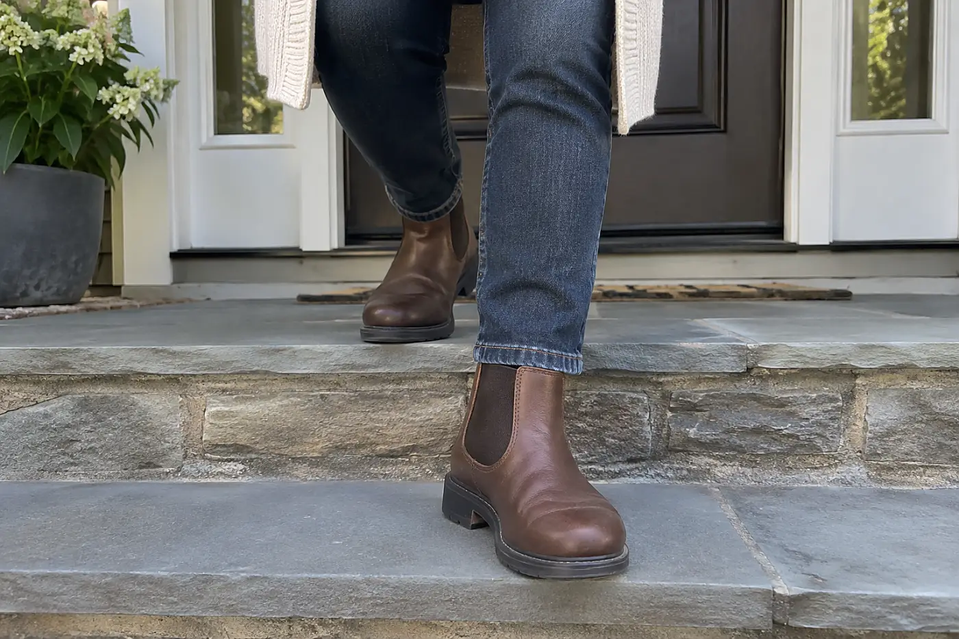 A Chelsea boot with a wide toe is centered on the foot of a woman over 50 stepping down her front stairs.