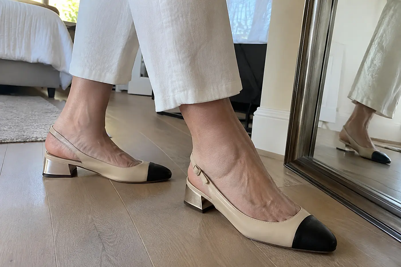 A slingback with a low heel is centered on the foot of a woman over 50 walking past a full-length mirror at home.