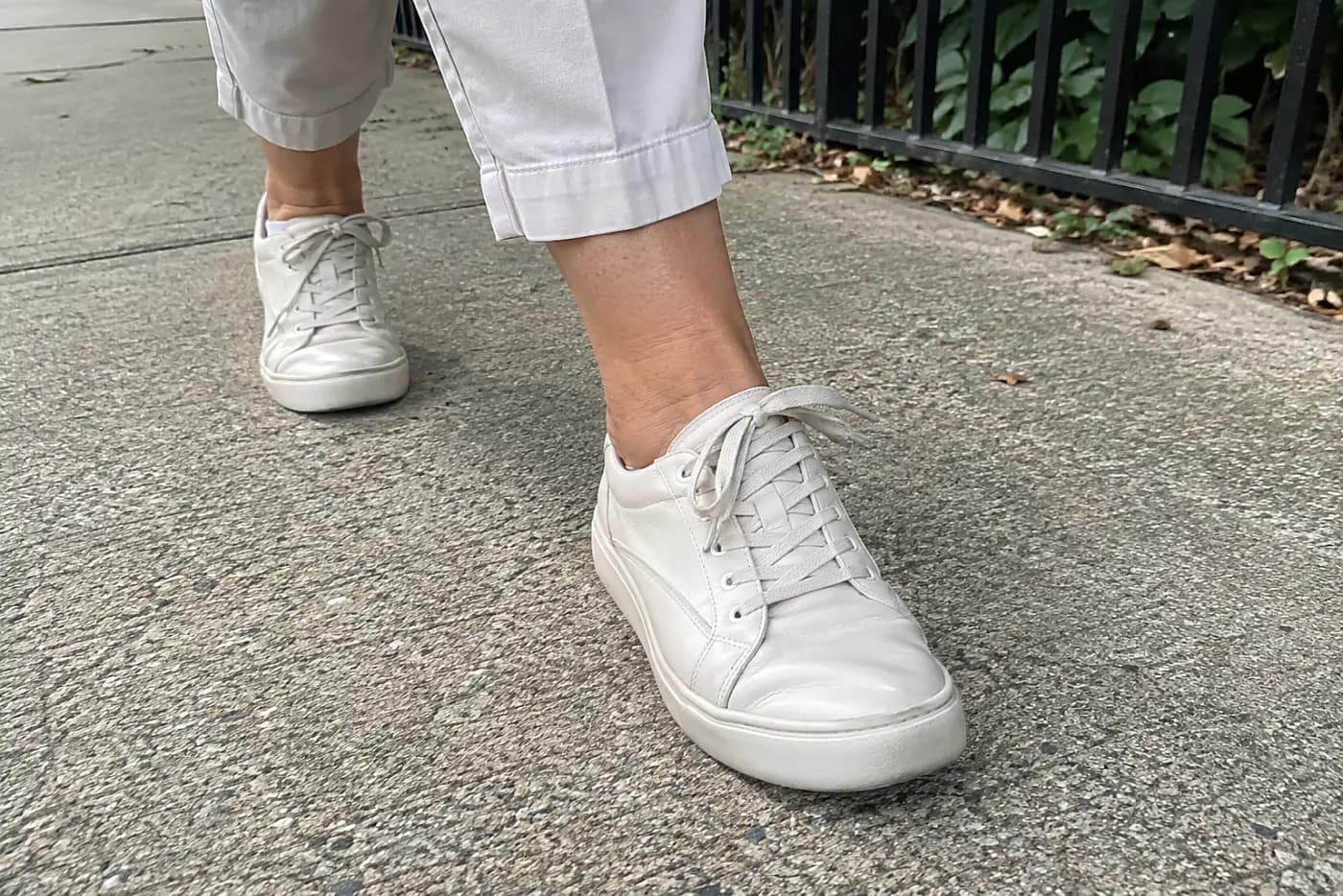 A soft leather sneaker is centered on the foot of a woman in her sixties stepping onto a sidewalk.