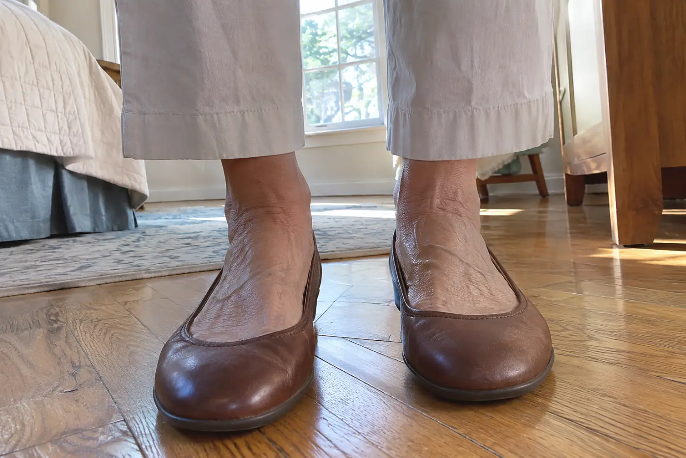 A wide-toe leather flat is centered beneath cropped pants on a woman over 50 standing with both feet flat on a wooden floor.