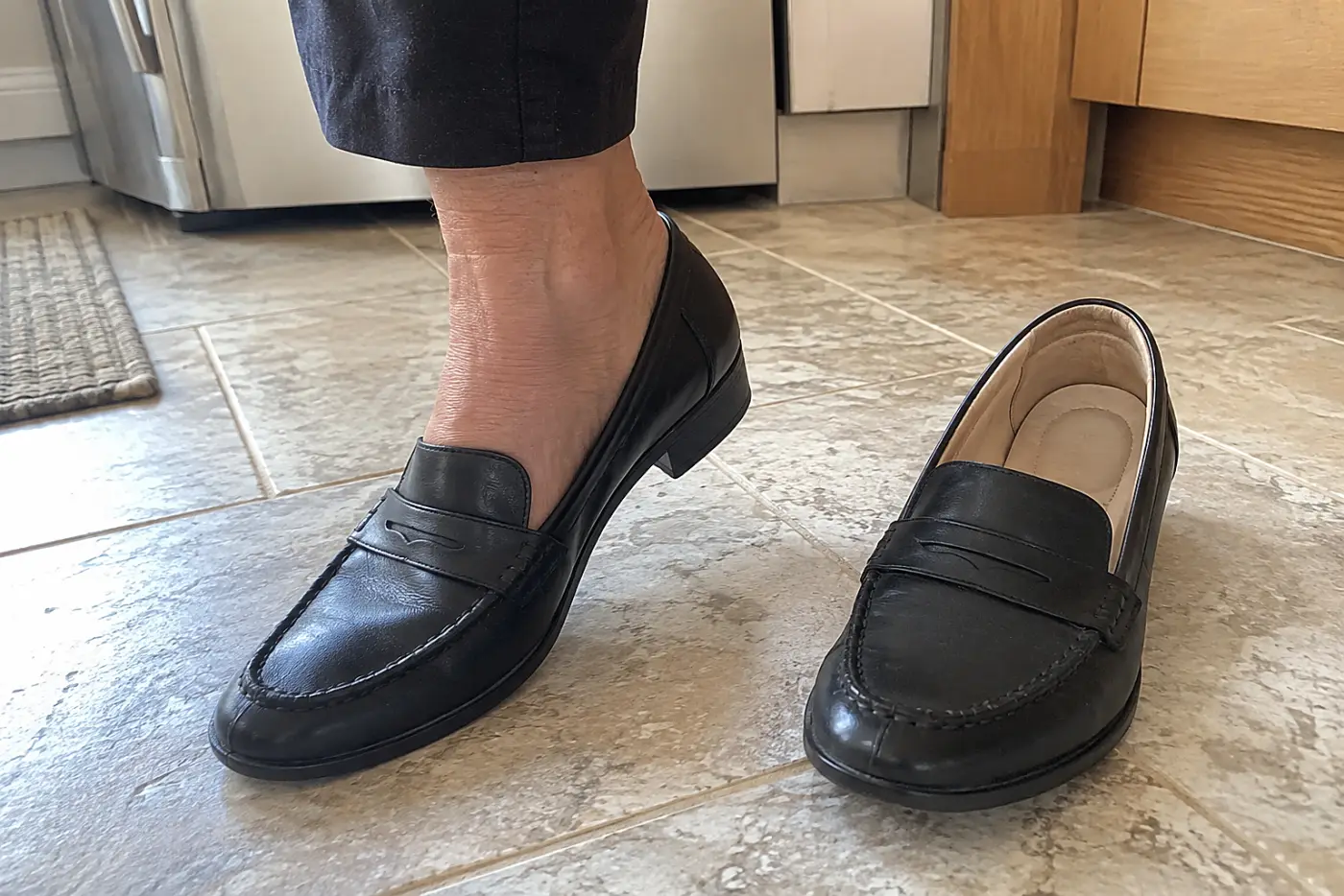 A leather loafer with cushioning is centered on the foot of a woman over 50 walking across a kitchen floor, with its padded interior visible in the matching shoe nearby.