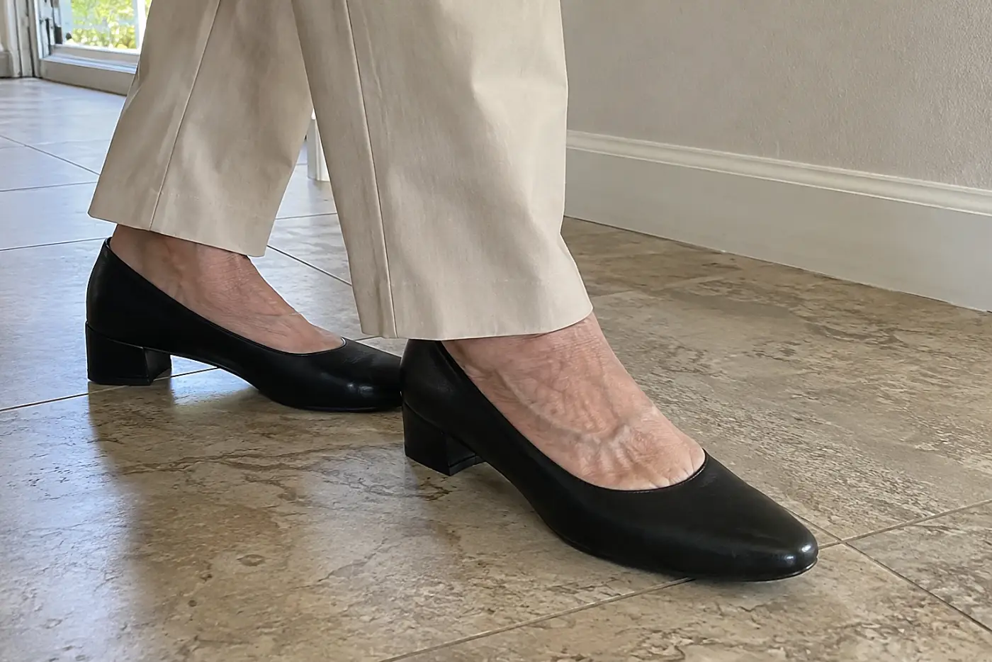 A low block-heel pump is centered beneath the trouser hem of a woman in her sixties standing in a hallway.
