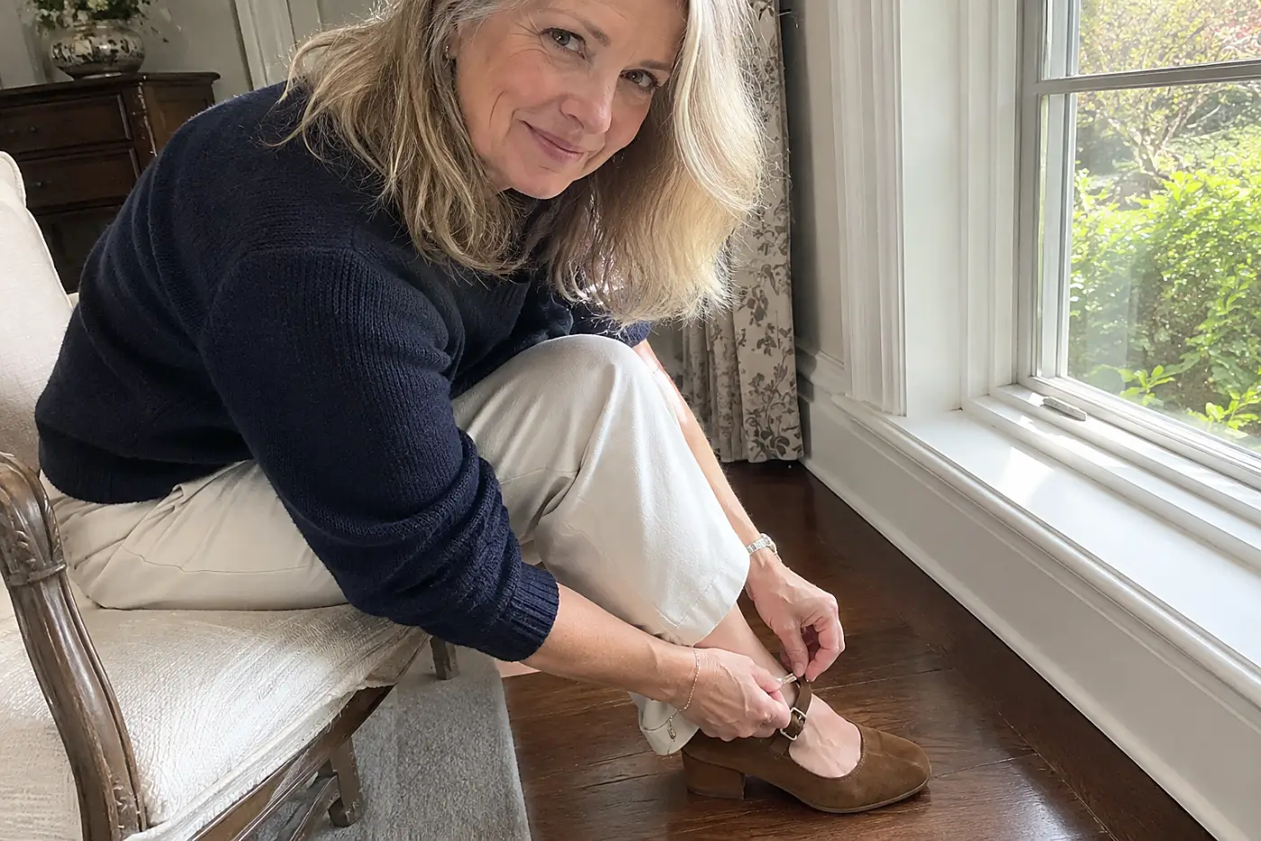 A suede Mary Jane is centered on the foot of a woman over 50 fastening its strap while seated by a window.