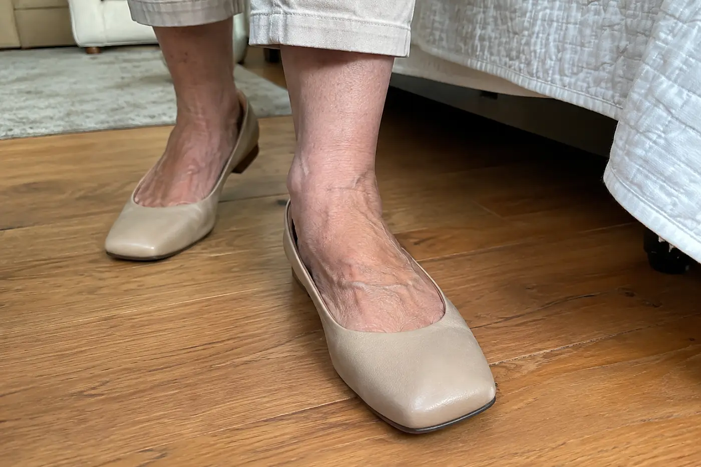 A square-toe ballet flat is centered on the foot of a woman in her sixties standing on a wooden floor beside her bed.