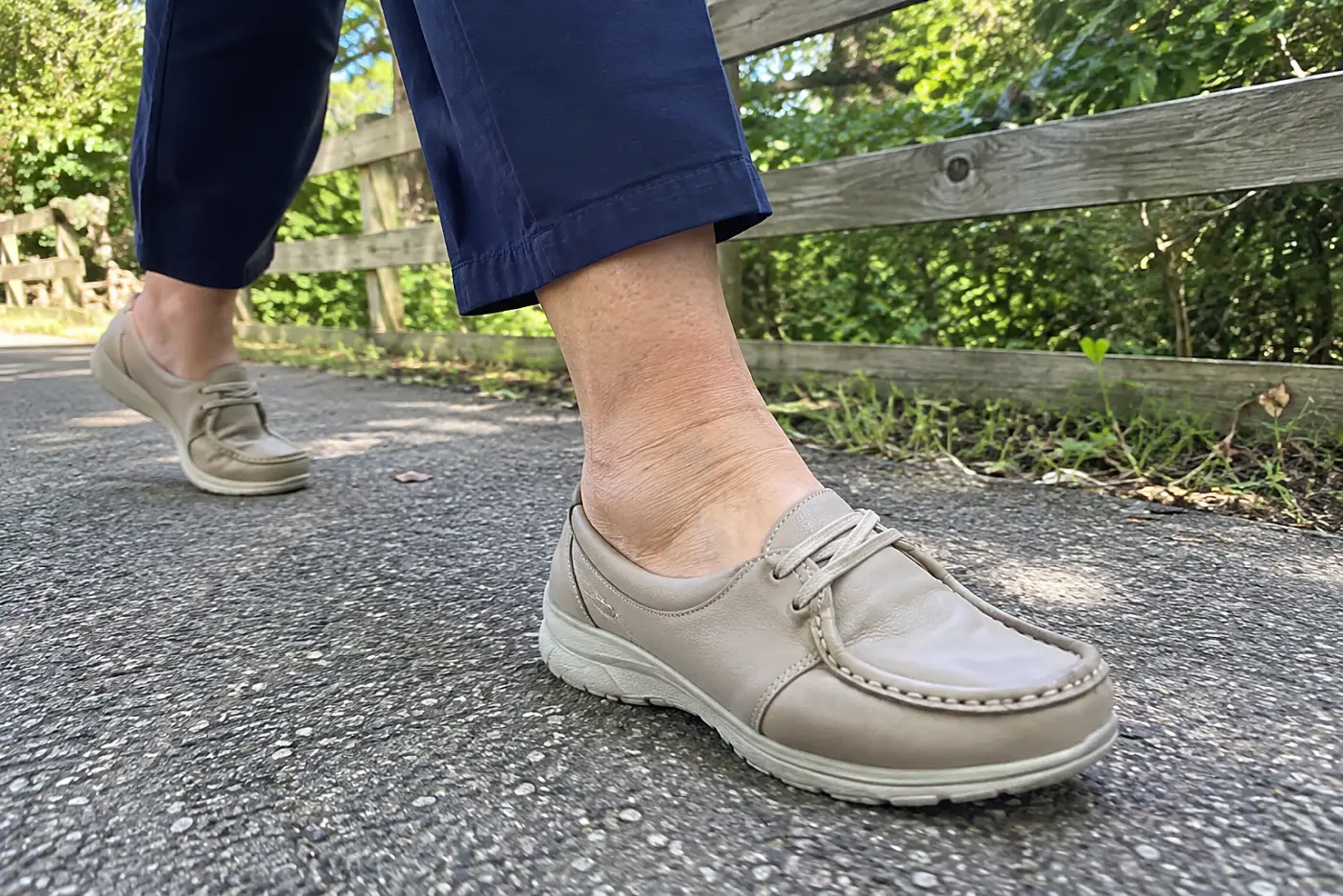 A moc-toe walking shoe is centered on the foot of a woman in her sixties taking a long step along a park path.