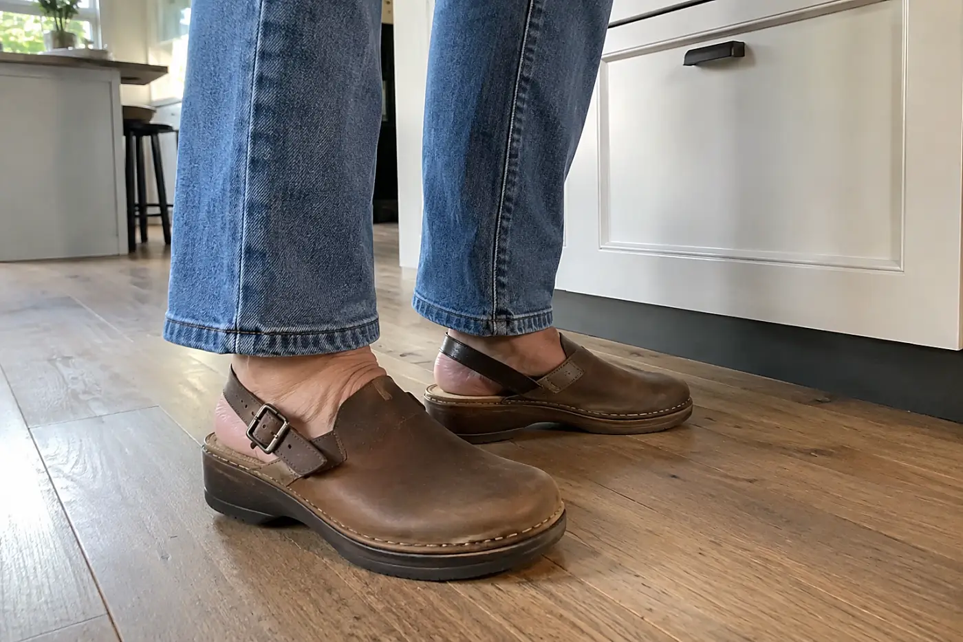 A buckled clog with a backstrap is centered on the foot of a woman over 50 standing beside a kitchen counter in straight jeans.