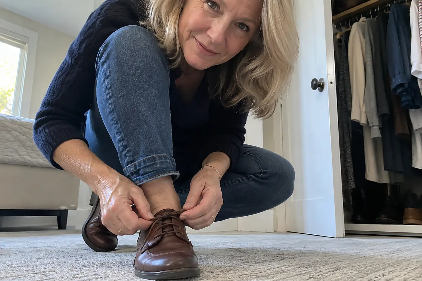 A lace-up derby shoe is centered on the foot of a woman over 50 fastening its laces beside her closet.