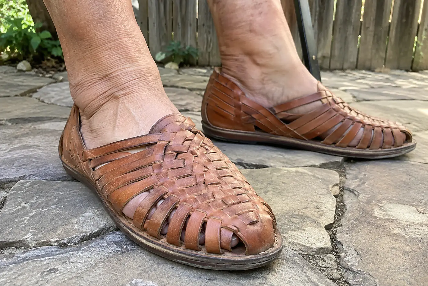 A leather huarache sandal is centered on the foot of a woman in her sixties sitting outside with both feet on a stone path.