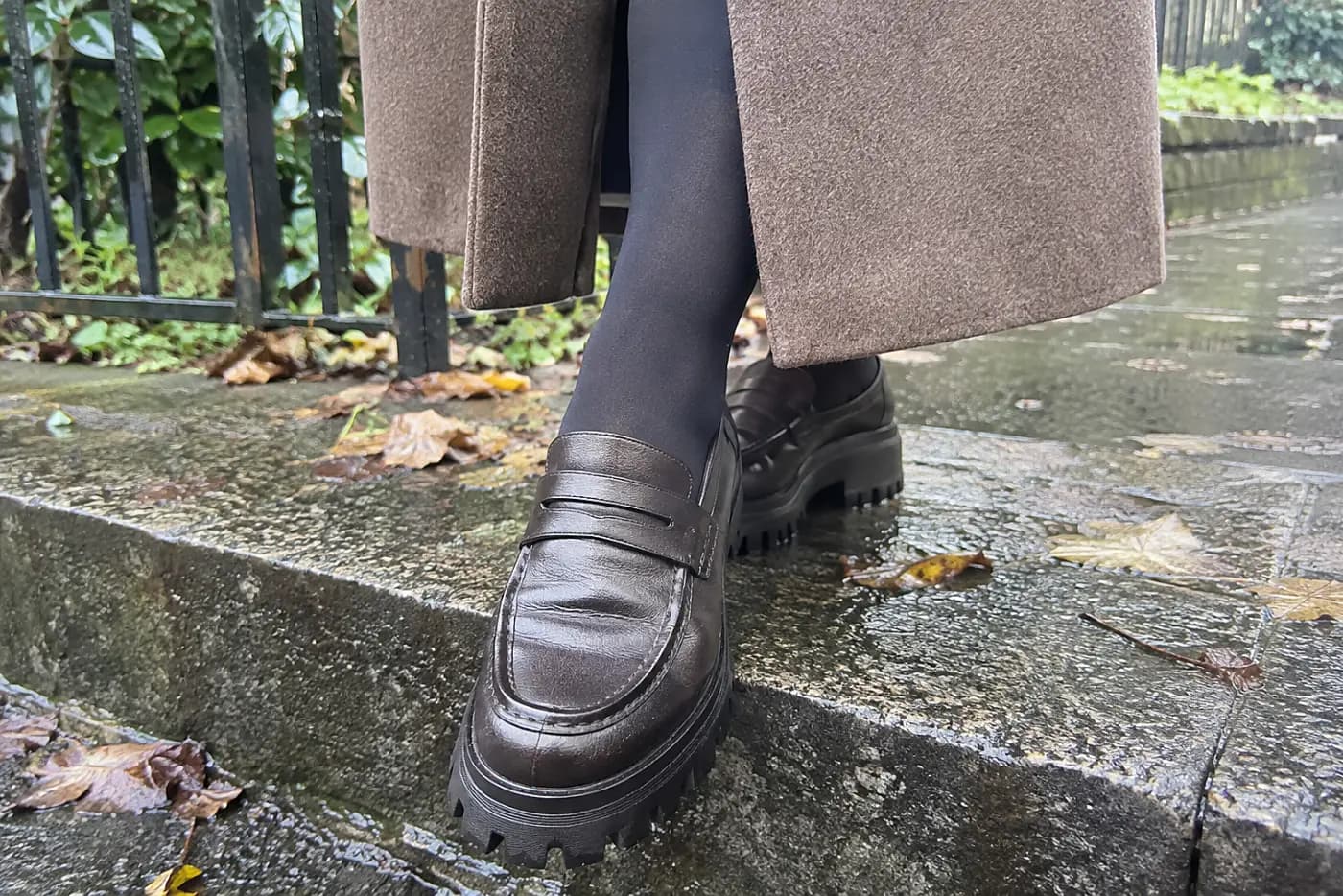 A lug-sole loafer is centered on the foot of a woman over 50 stepping off a damp curb in a long coat.