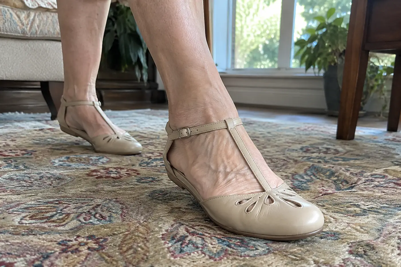 A T-strap flat is centered on the foot of a woman in her sixties walking across a living-room rug.