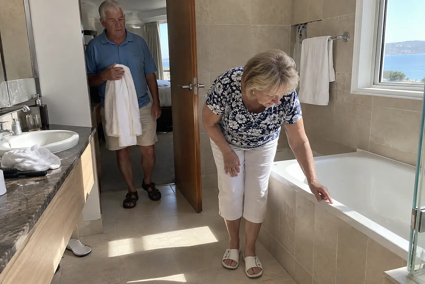 Older woman looking at the edge of a bathtub in a hotel bathroom.