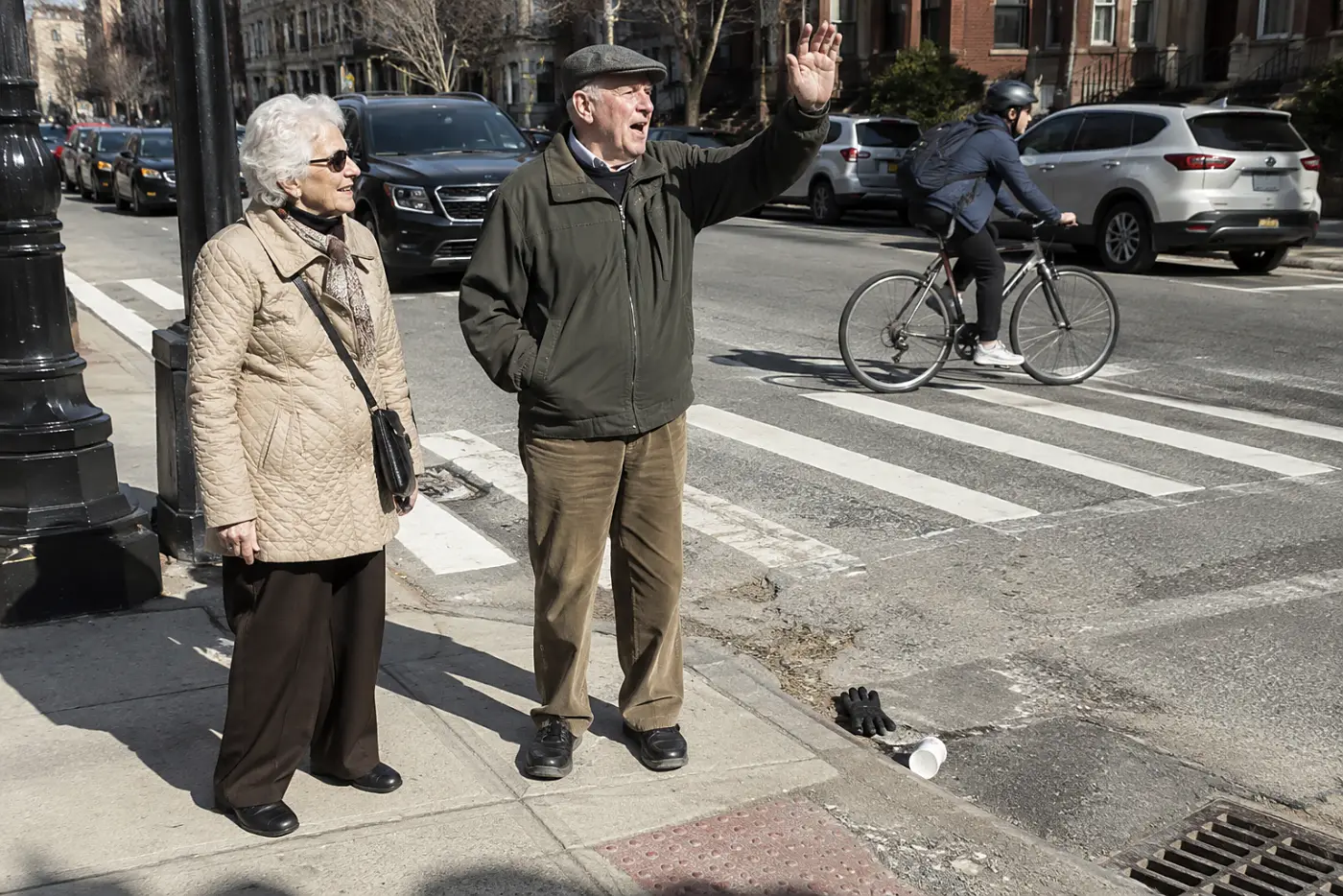 Older woman waiting beside a companion at a crosswalk on a busy street.