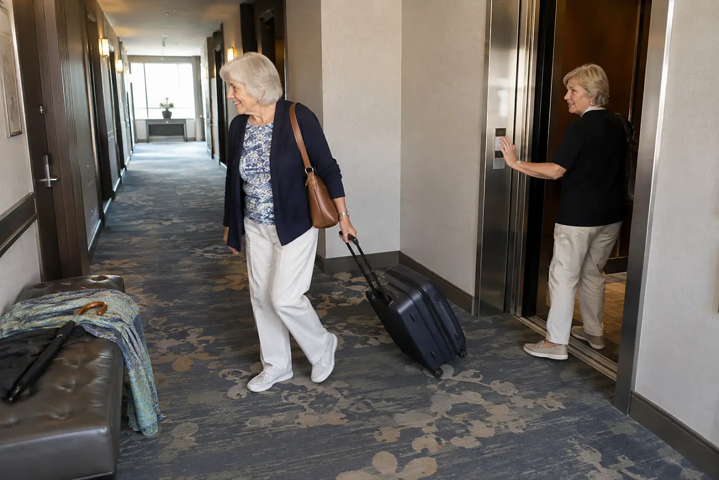 Older woman looking from an elevator toward her room door in a hotel hallway.