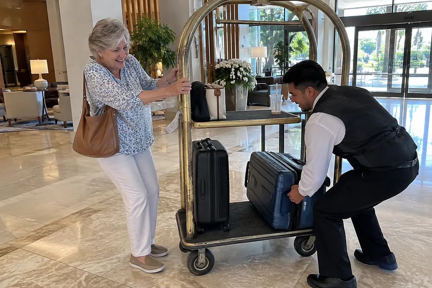 Older woman standing beside a luggage cart while a hotel employee lifts a suitcase in the lobby.