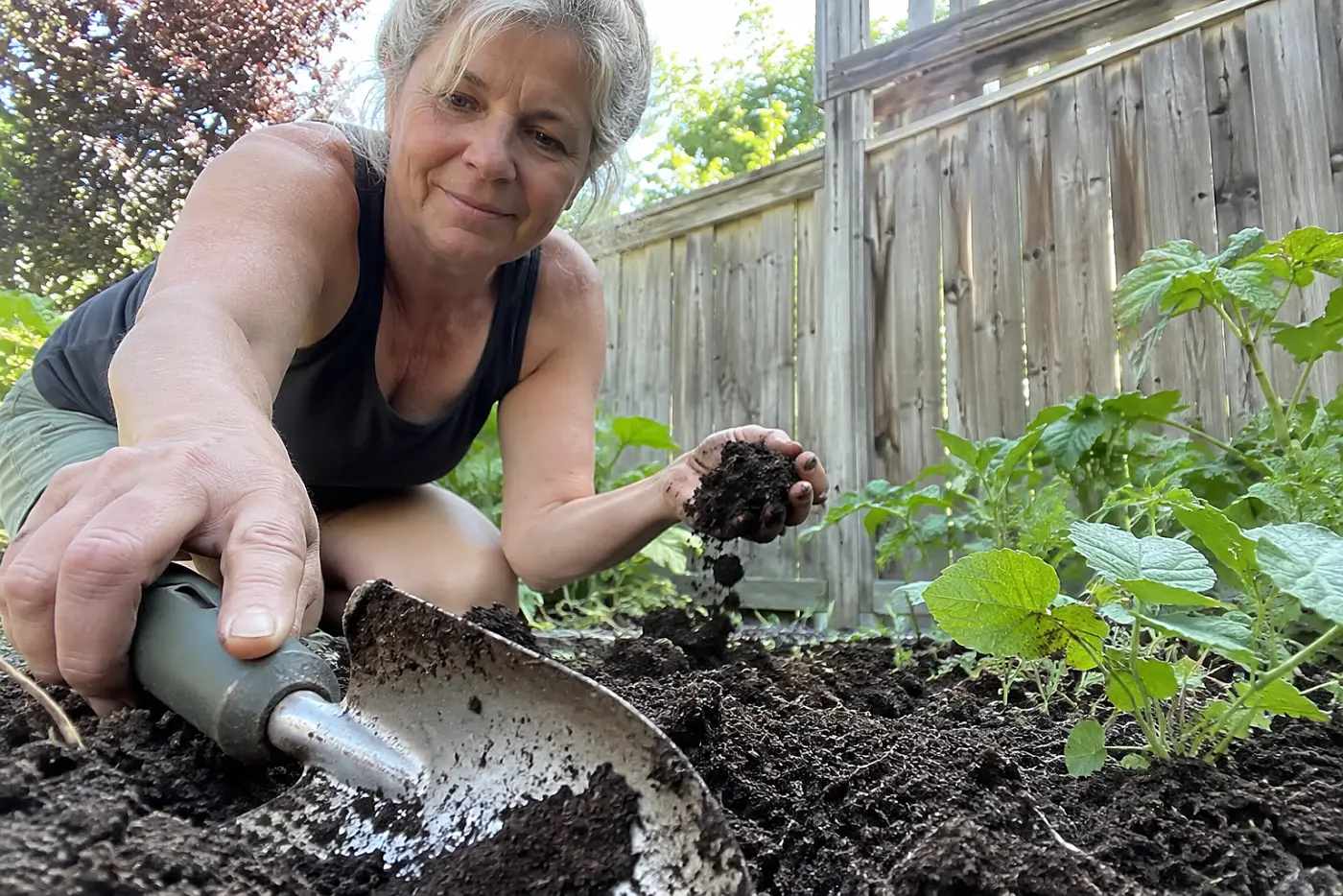 A trowel beside the bed lies next to fresh soil in a backyard garden, with a woman over 50 working nearby and no visible writing.