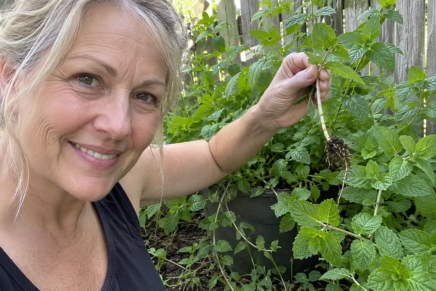 Mint beyond its pot reaches into a nearby garden bed, with a woman over 50 lifting a runner and no visible writing.