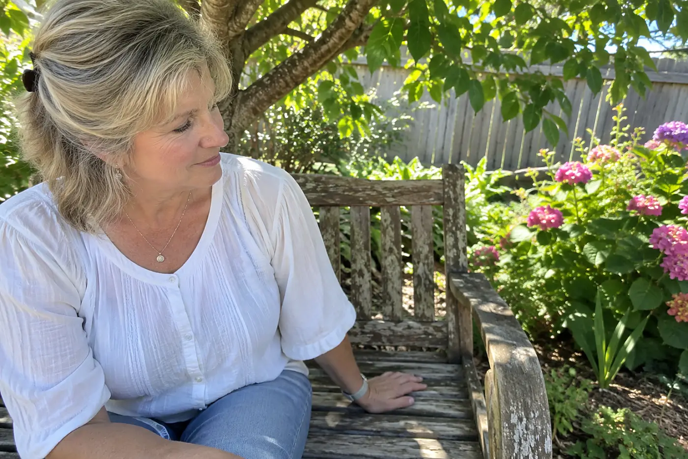 A bench under the tree faces backyard flowerbeds, with a woman over 50 sitting on it and no visible writing.