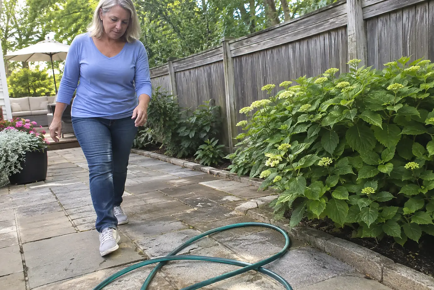 A hose across the path lies in a loose coil on backyard paving, with a woman over 50 approaching it and no visible writing.