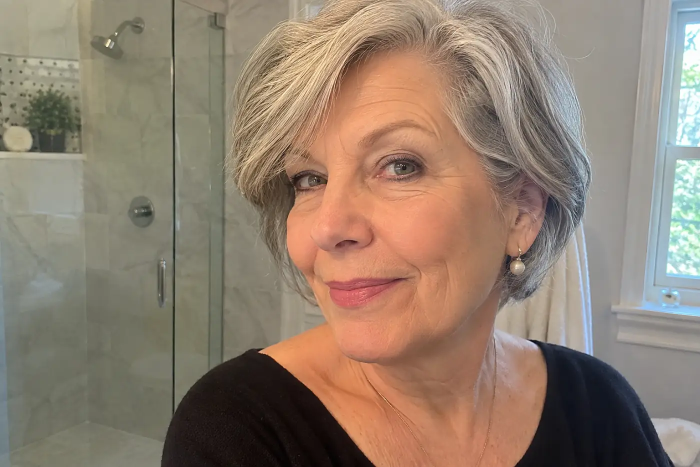 A woman in her sixties makes her final bathroom mirror check under warm bulbs before turning toward a nearby window, with no writing visible.