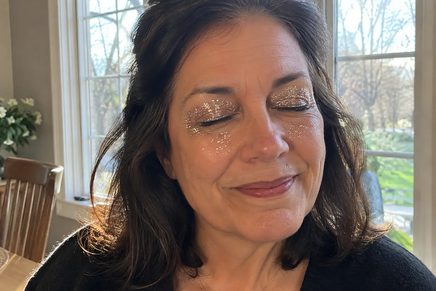 Glitter across both eyelids and scattered onto the cheeks fills the frame on a woman in her fifties at her dining room table in daylight, with no writing visible.