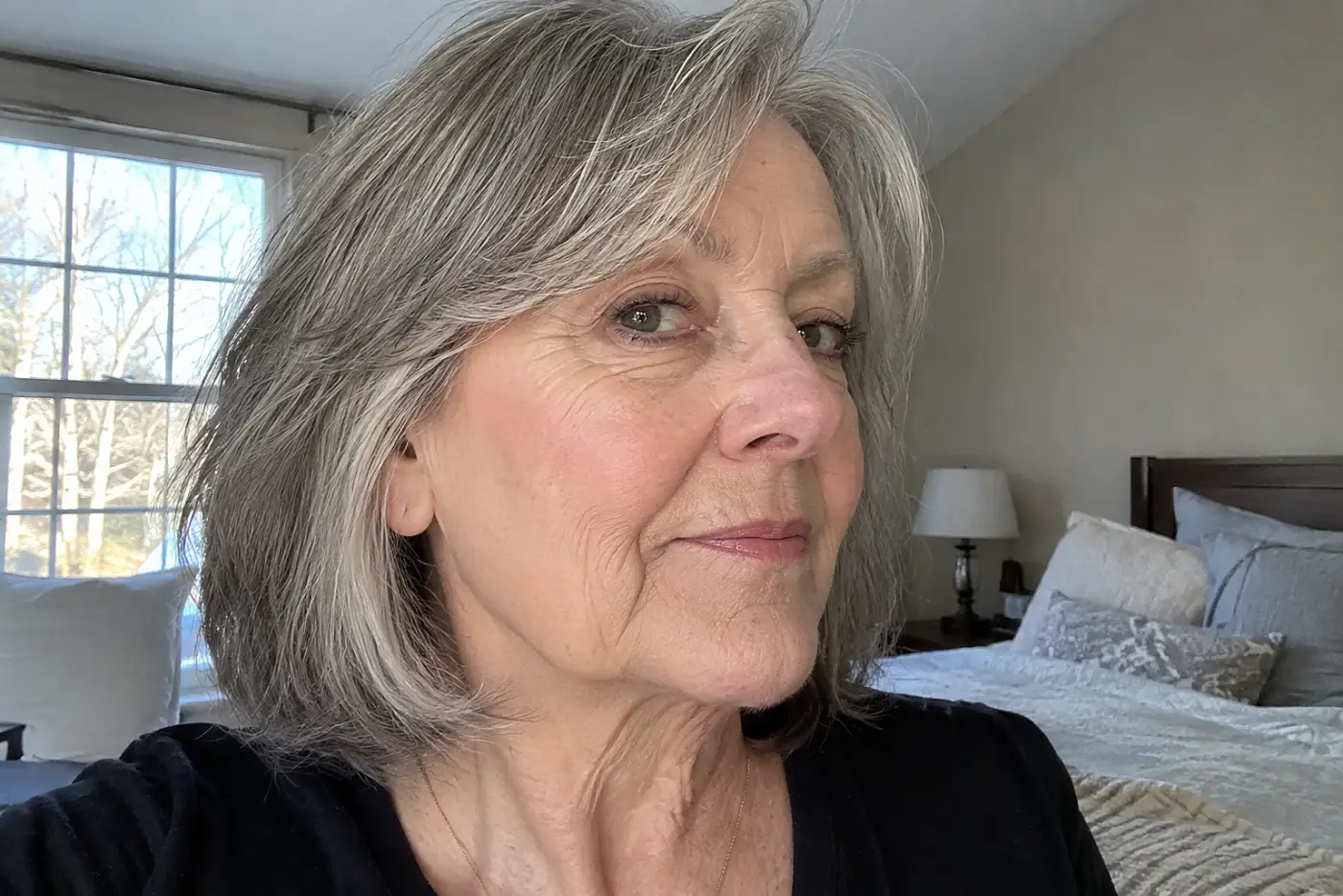 Blush placed low on the cheeks is centered on a woman in her sixties checking a mirror in her bedroom, with no writing visible.