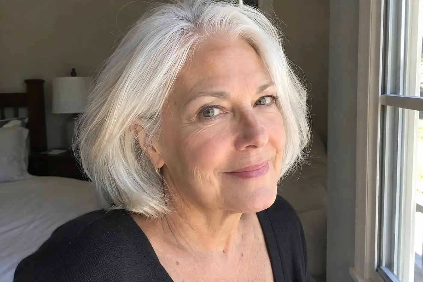 A frosty brow bone stripe catches daylight on a woman in her sixties standing beside her bedroom window, with no writing visible.