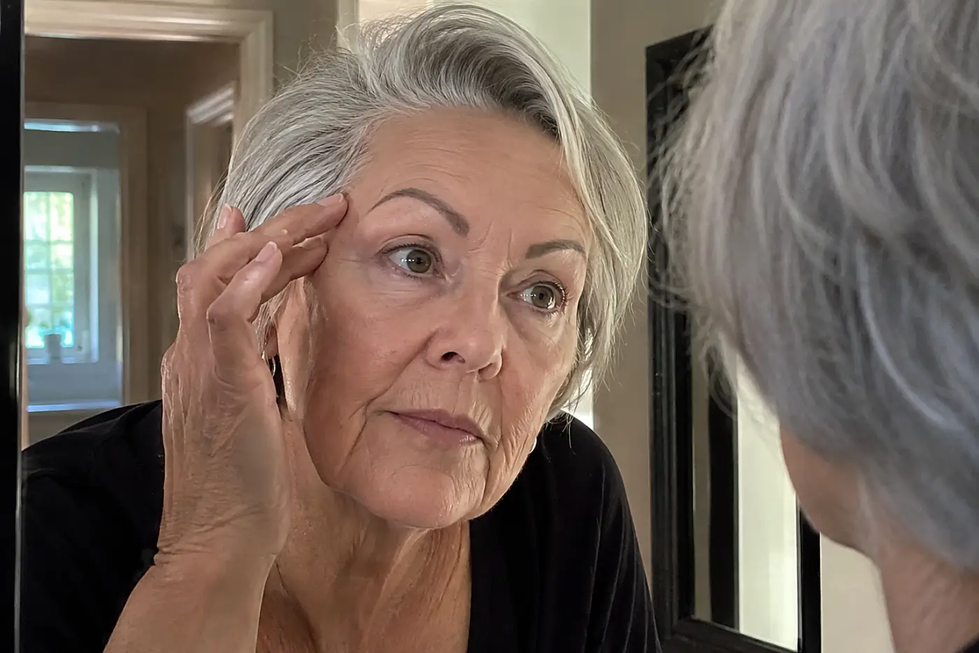 A high pencil brow arch is centered on a woman in her sixties leaning toward her hallway mirror, with no writing visible.
