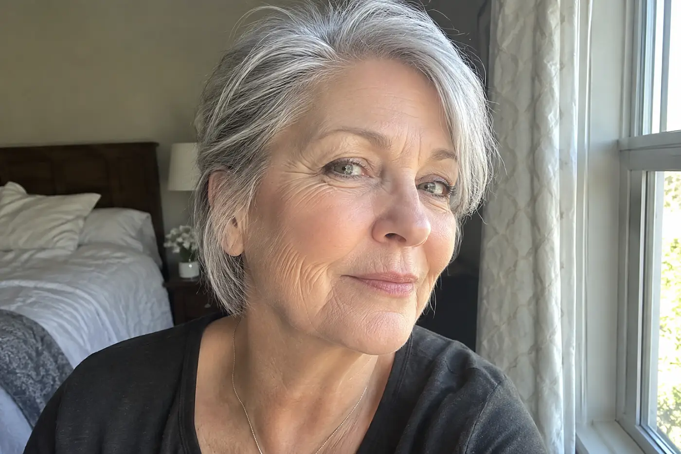 Matte foundation across the cheeks is centered on a woman in her sixties standing beside a bedroom window, with no writing visible.