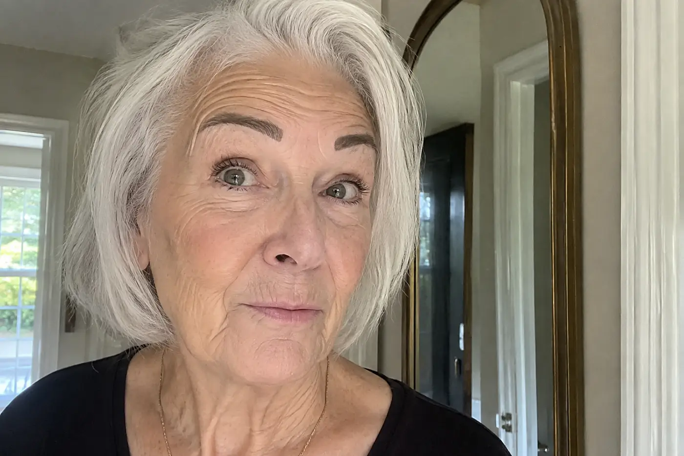 Heavy powder brows are centered on a woman in her sixties checking her reflection in a hallway mirror, with no writing visible.
