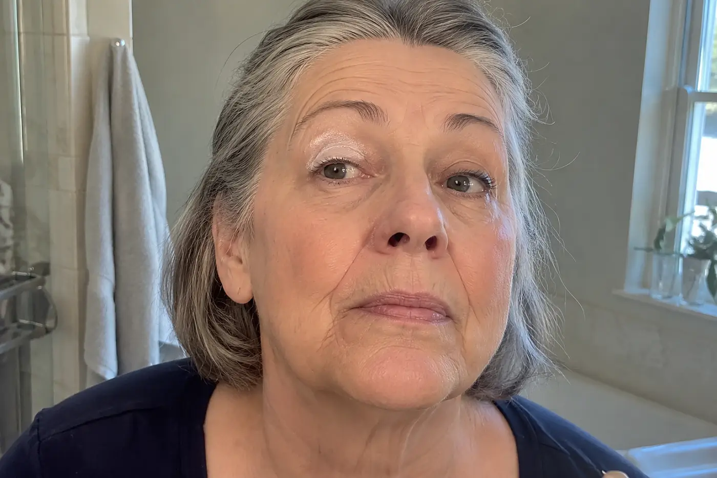 Unblended cream shadow on one lid is centered on a woman in her sixties looking up into her bathroom mirror, with no writing visible.