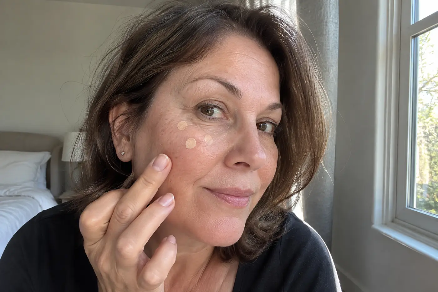 Concealer dabs over freckles are centered on a woman in her fifties sitting near a bedroom window, with no writing visible.