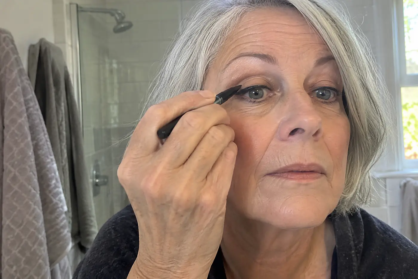 A thick line above the upper lashes is centered on the open eye of a woman in her sixties at her bathroom mirror, with no writing visible.