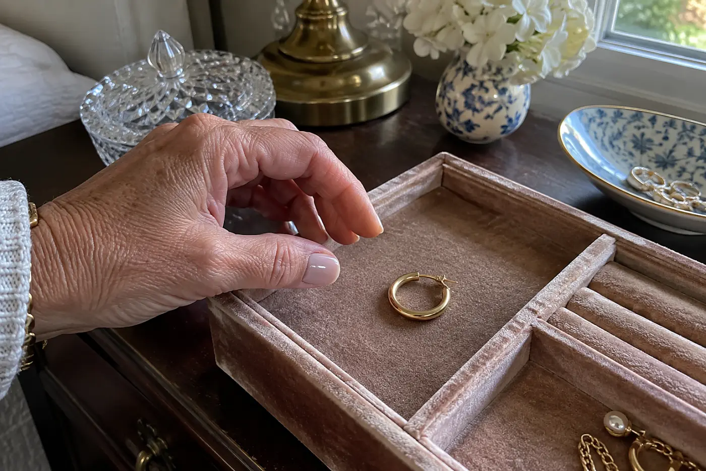 A single gold hoop earring lies in the center of a worn velvet jewelry tray on a bedroom dresser, beside the hand of a woman in her sixties.